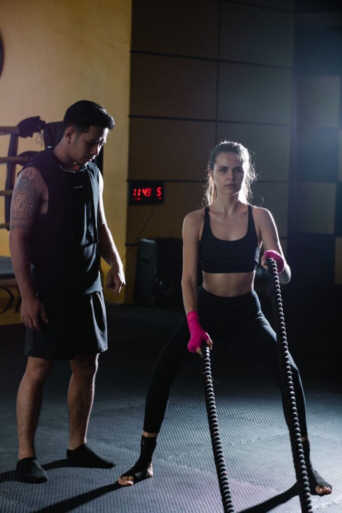 Full body Latin American personal instructor helping active female in activewear exercising with battle ropes during intense training in gym